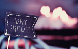 closeup of a black flag-shaped signboard with the text happy birthday, and a birthday cake with lit candles in the background